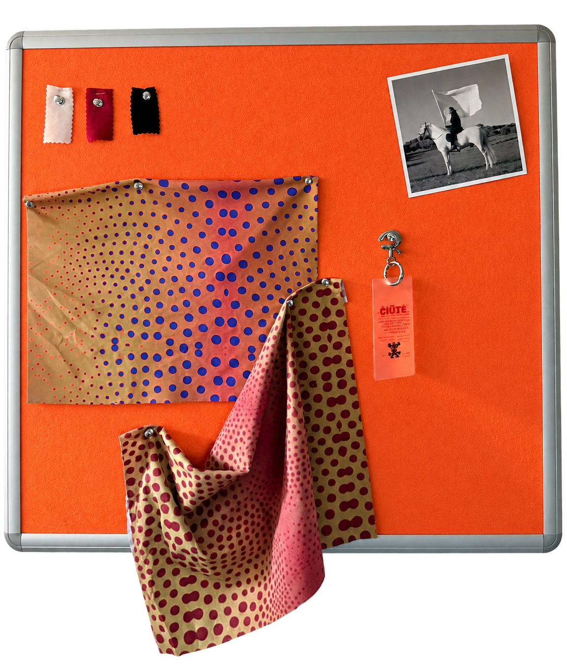 An orange fabric bulletin board with a silver metal frame, styled with three fabric swatches, a black-and-white photo print, a small keychain, and two draped patterned scarves.
